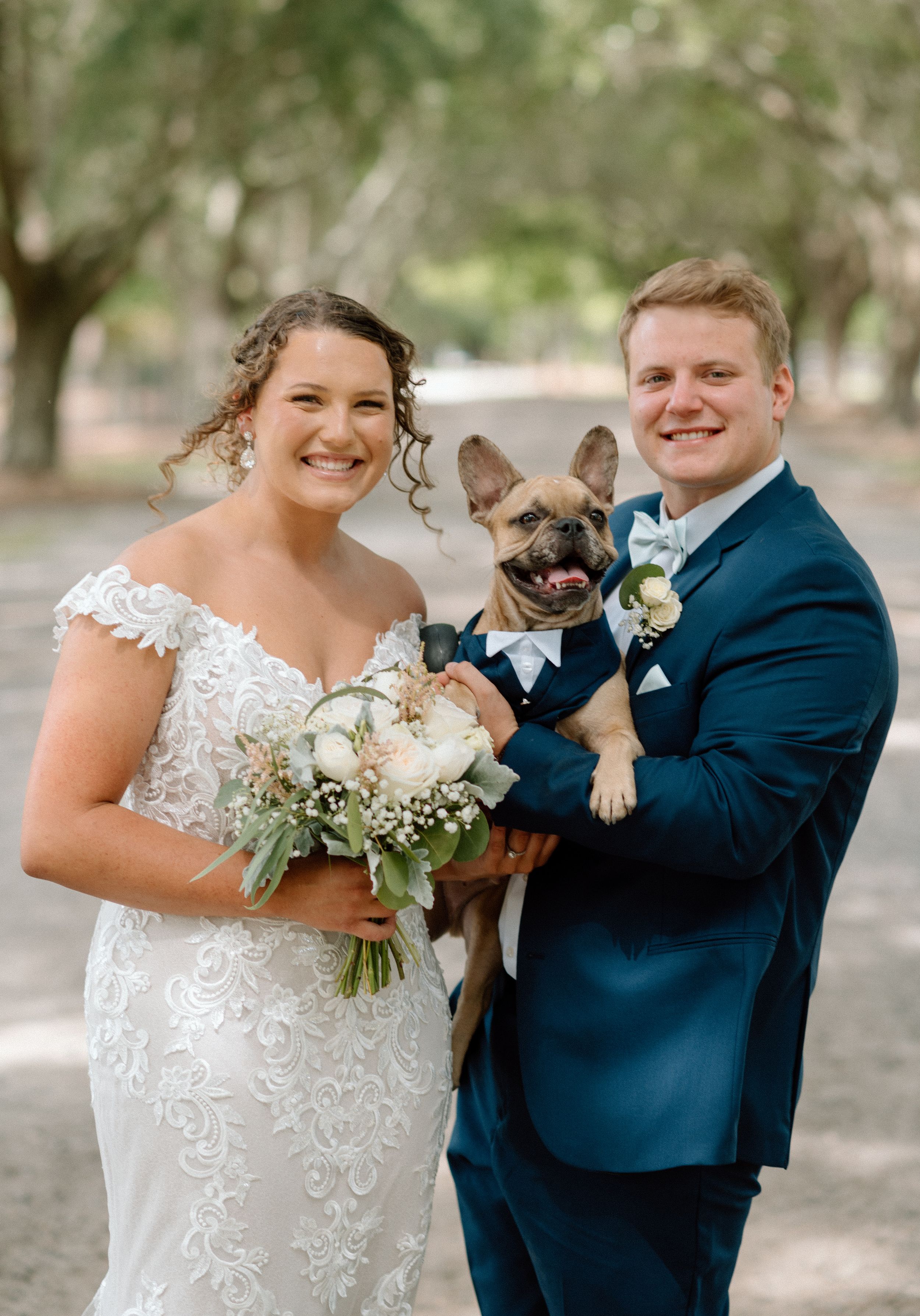 A wedding couple poses with their French Bulldog wearing a bow tie.