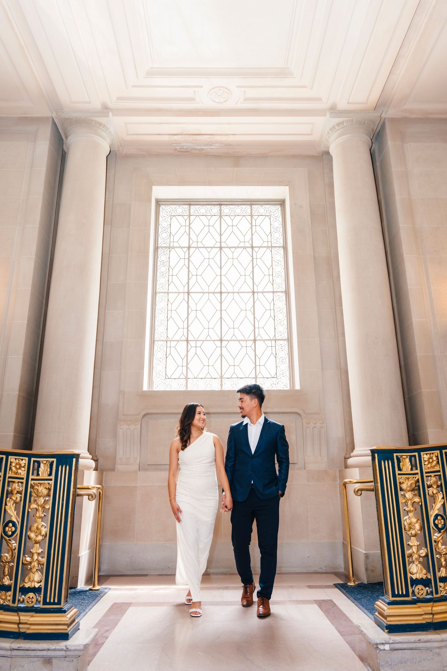 Elegant couple stands in ornate hallway with tall window and decorative railings in grand building.