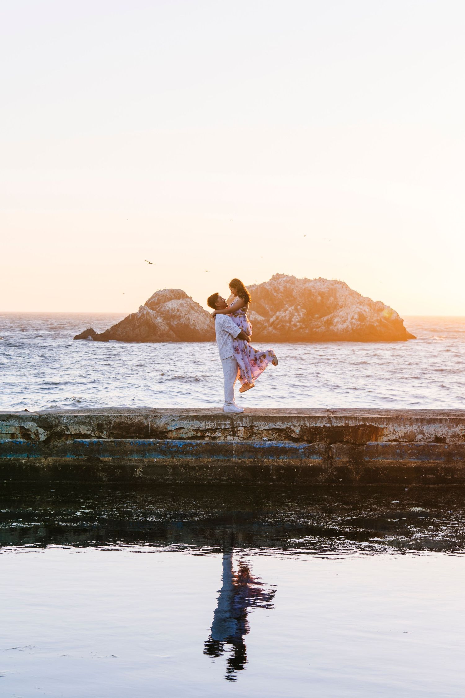 Couple embracing at sunset by rocky coastline with reflection in water.