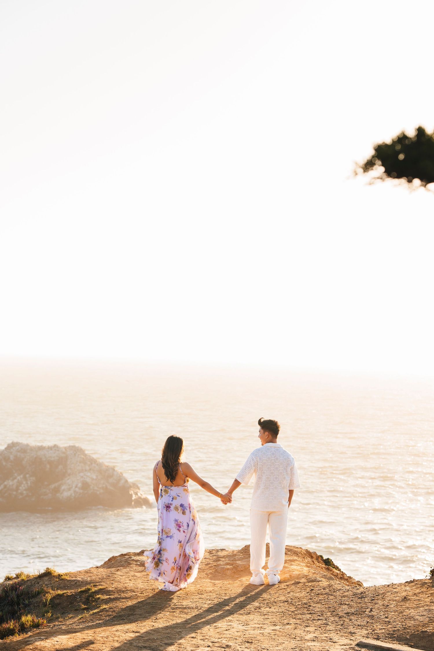 Couple holding hands while standing on coastal cliff during sunset.