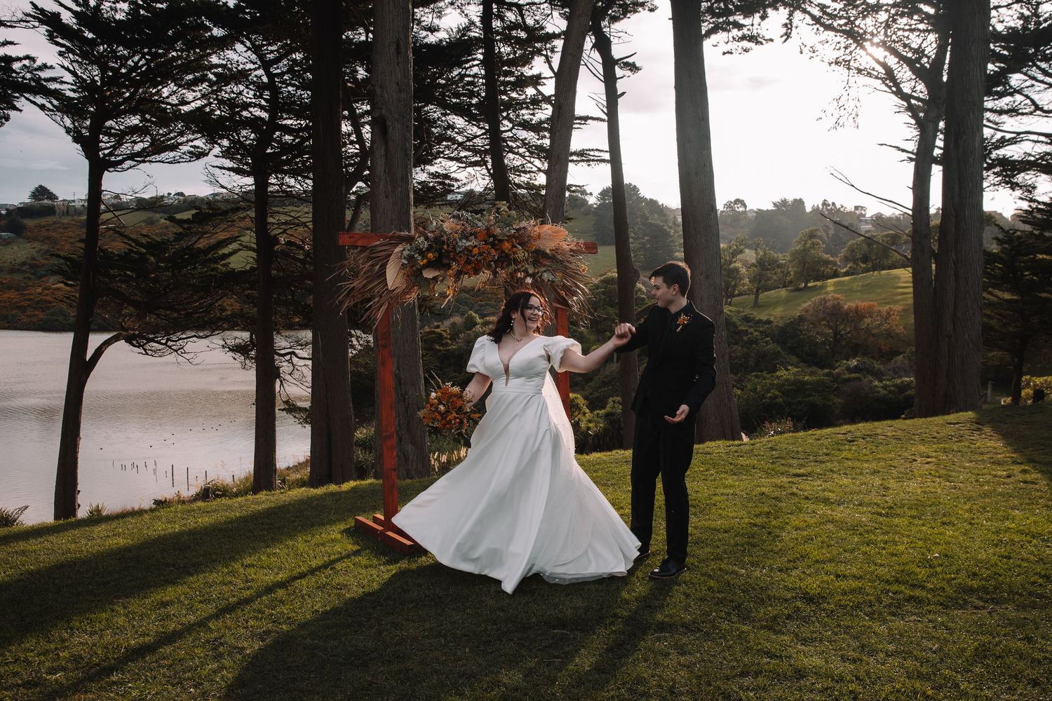 A joyful wedding dance moment captured by a lake with tall pine trees.