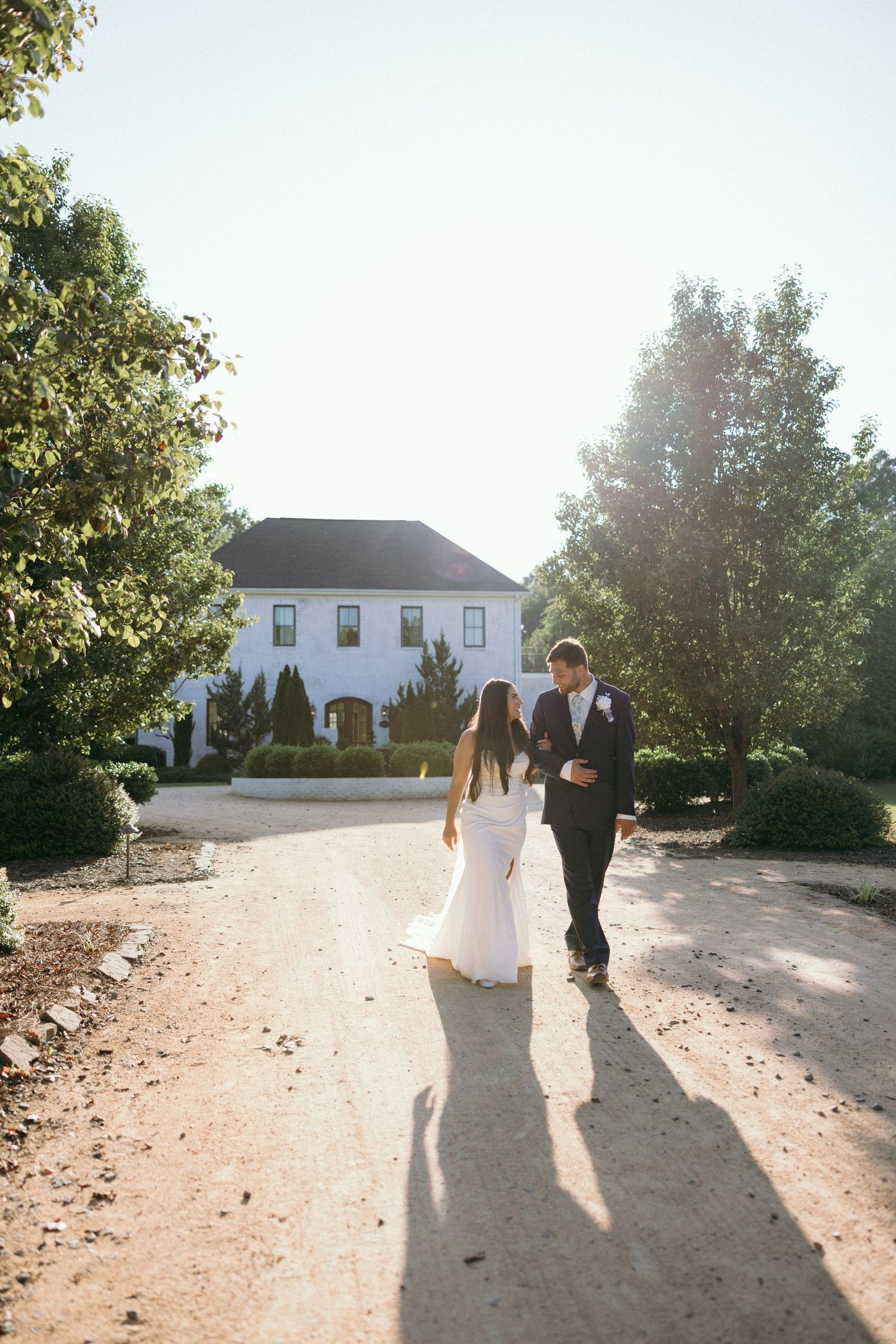 Couple walking down sunny path towards white building with beautiful landscaping. The Bradford, Separk  Mansion, Graylyn Estate