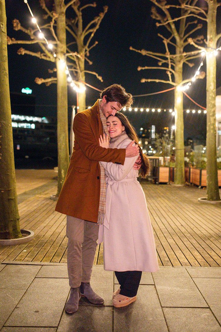 A happy couple hugs on an illuminated wooden boardwalk at night with string lights glowing in the trees above.
