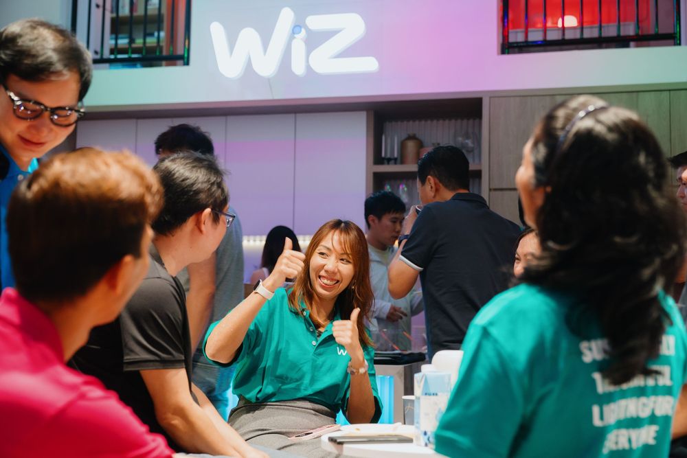 A group of people laughing and socializing at a cafe table under a neon Wiz sign.