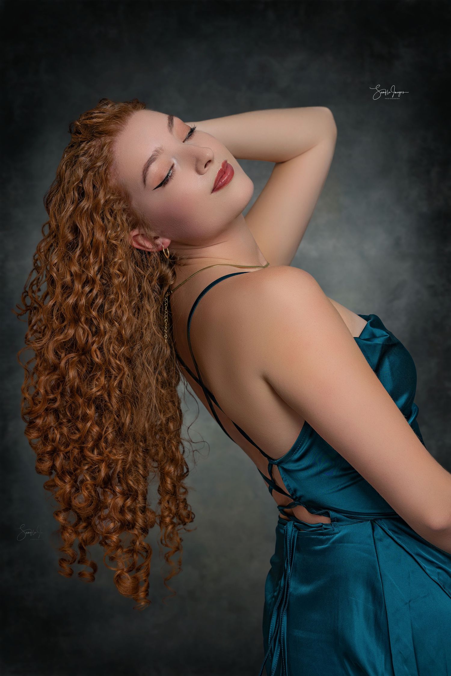A dramatic portrait featuring long curly red hair and a teal silk dress against a dark moody background.