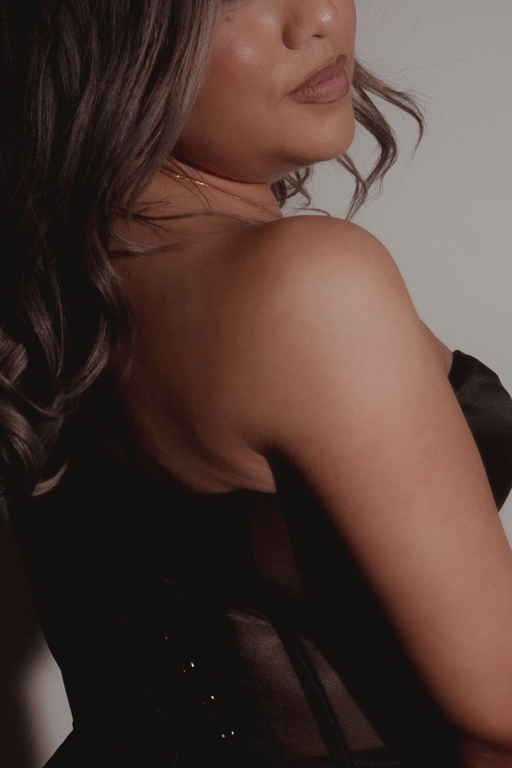 Close-up portrait showing bare shoulder and wavy brown hair against neutral background in moody lighting.