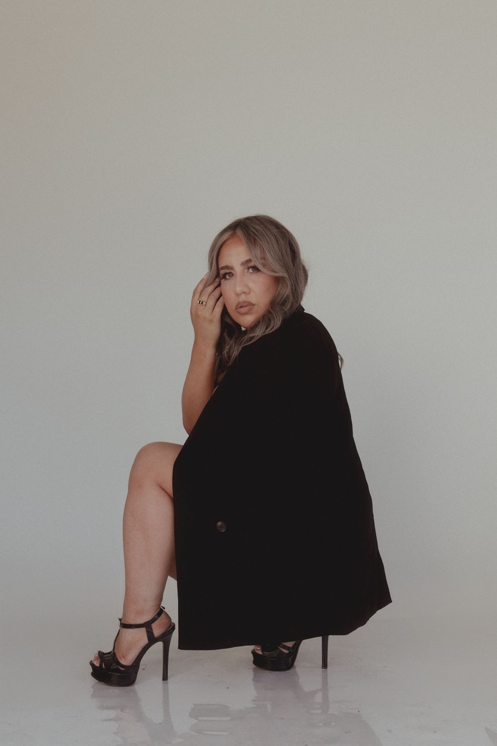 Fashion portrait of person in oversized black blazer and heels crouching against white background.