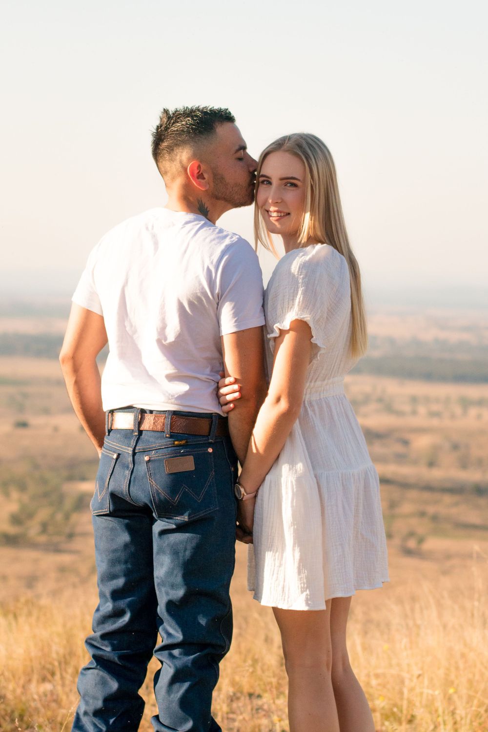 Couple in white outfits embracing on hilltop during sunset with scenic landscape behind them.
