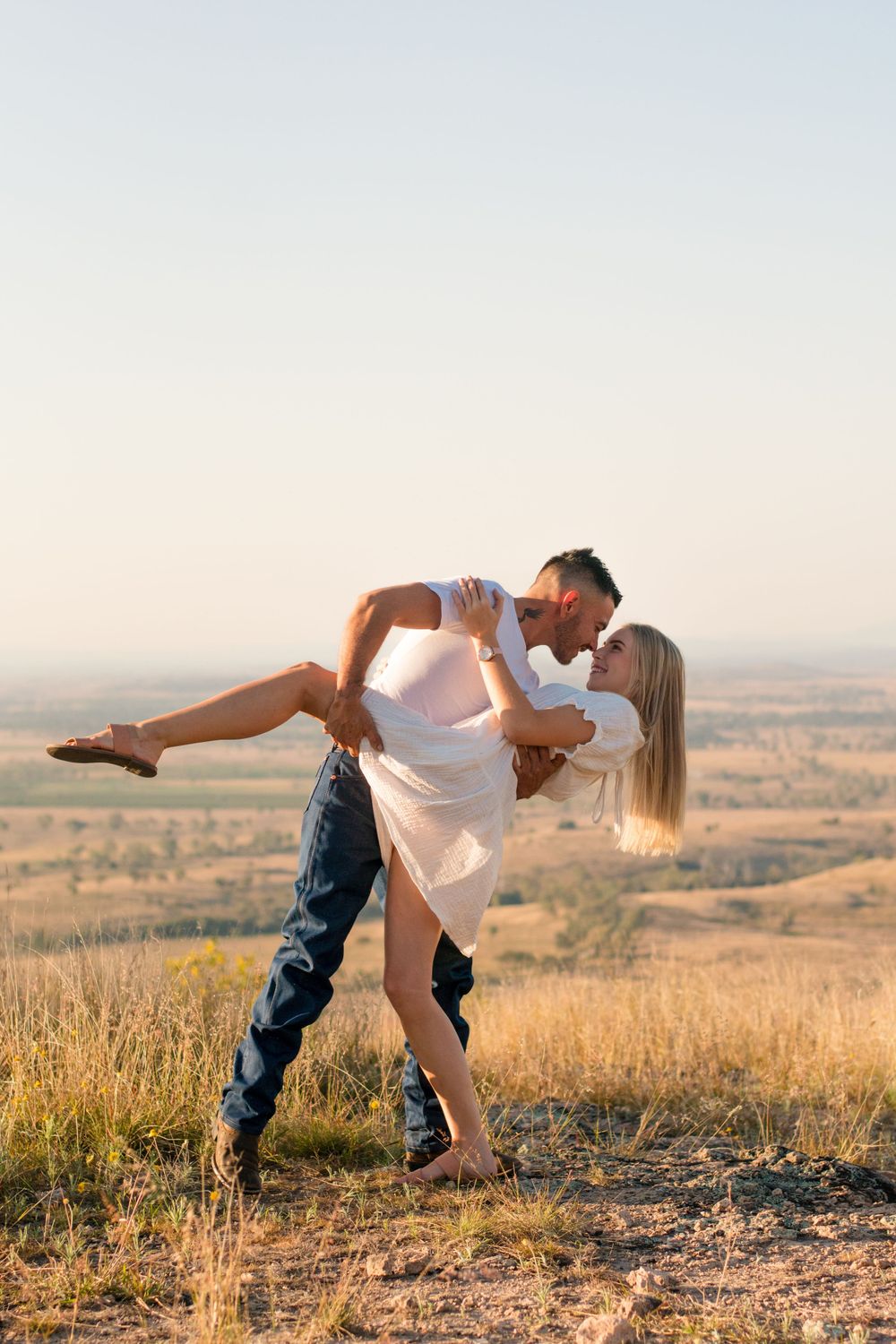 Romantic couple in casual attire embrace and dance on a hilltop overlooking a vast landscape at sunset.
