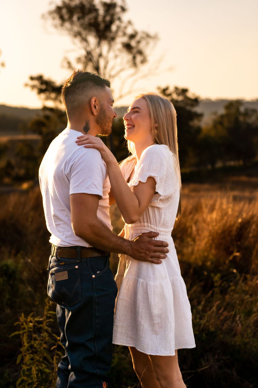 A couple shares a tender moment of laughter against a warm sunset backdrop in nature.