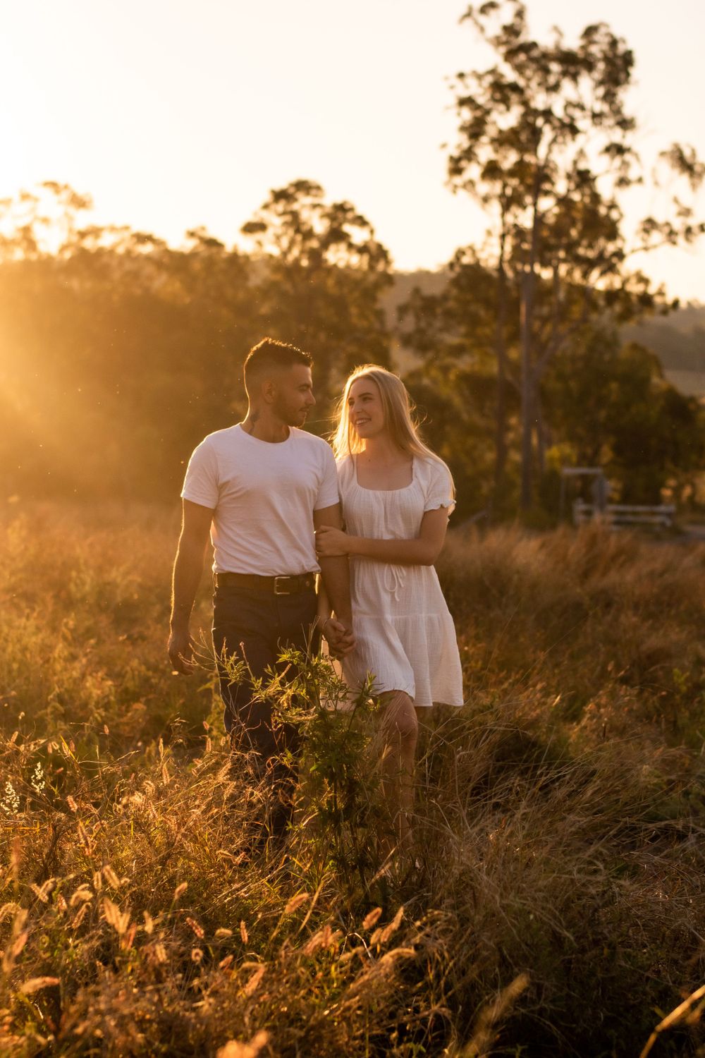 A couple walks together through golden grass during sunset in a scenic outdoor setting.