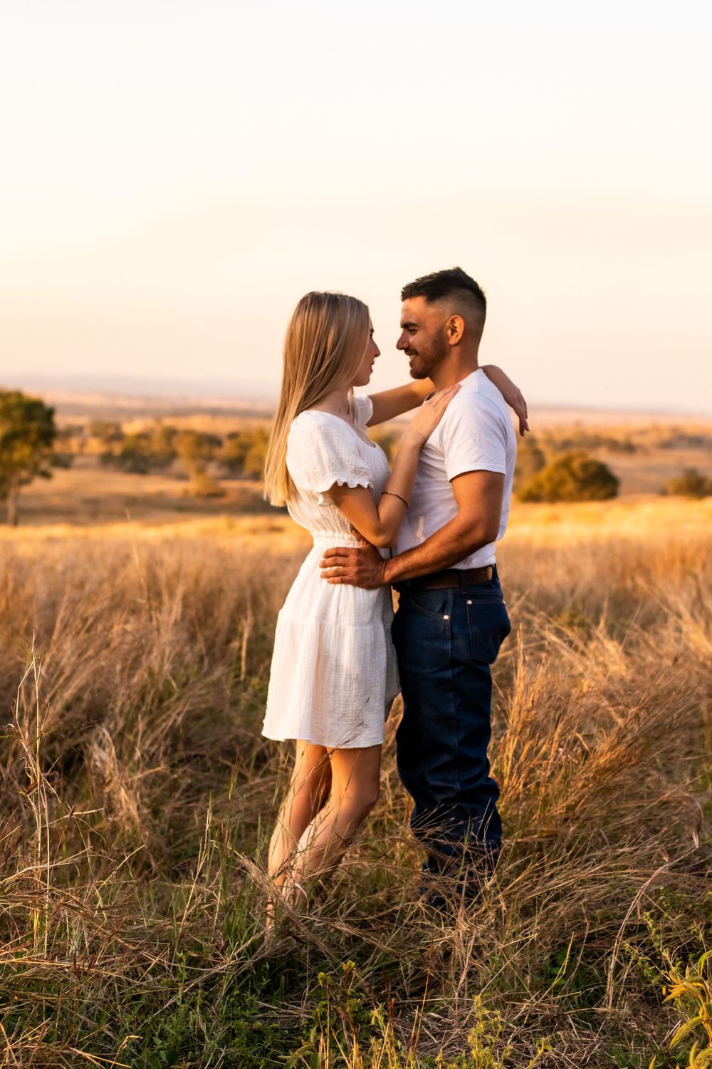 Couple sharing an intimate moment in white summer outfits against a golden sunset landscape.