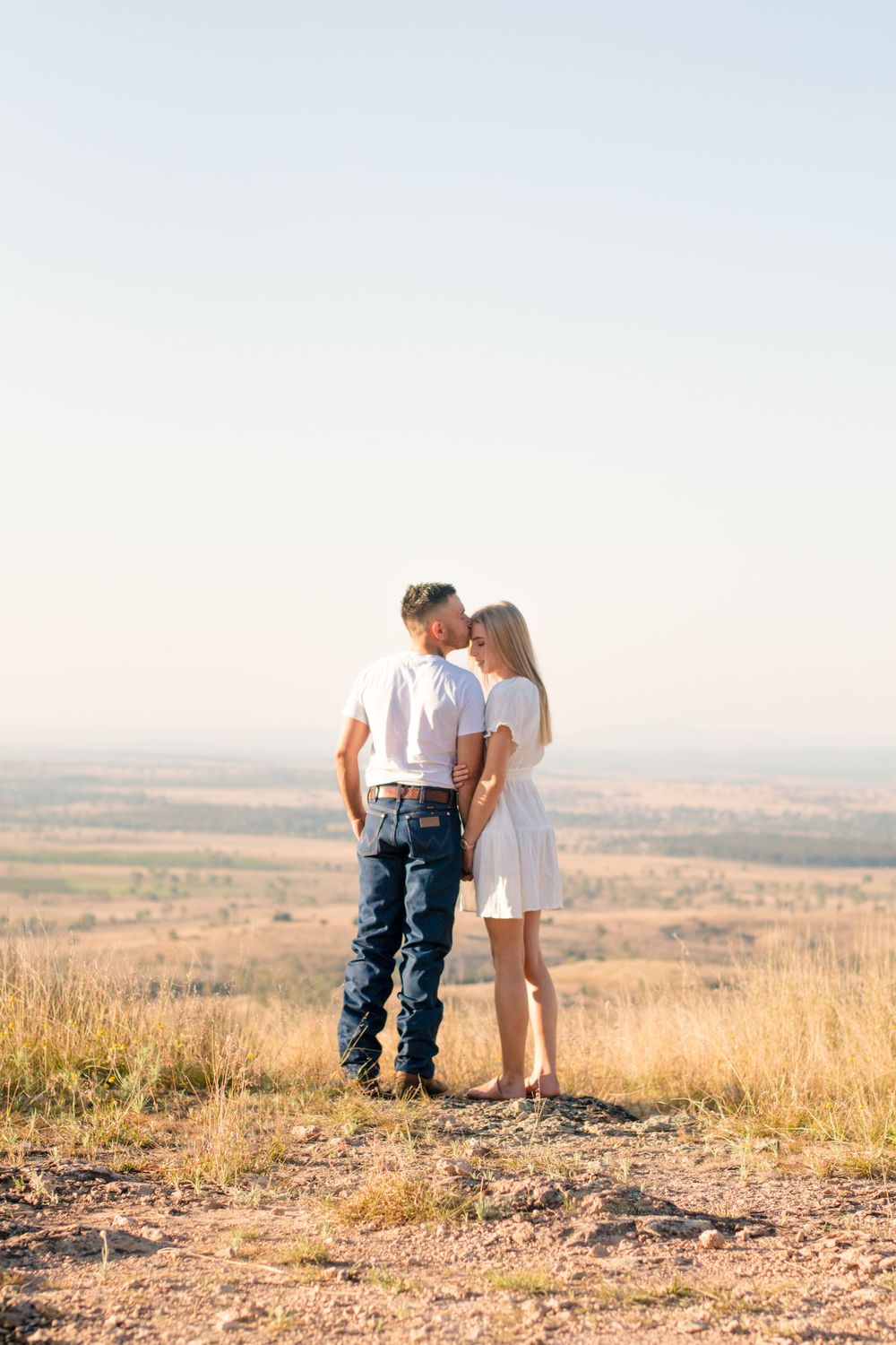 Couple stands close together on a hilltop overlooking a vast countryside landscape.
