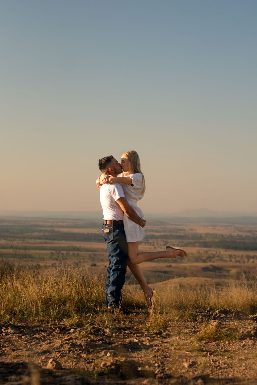 Romantic embrace on hilltop overlooking vast plains during sunset.