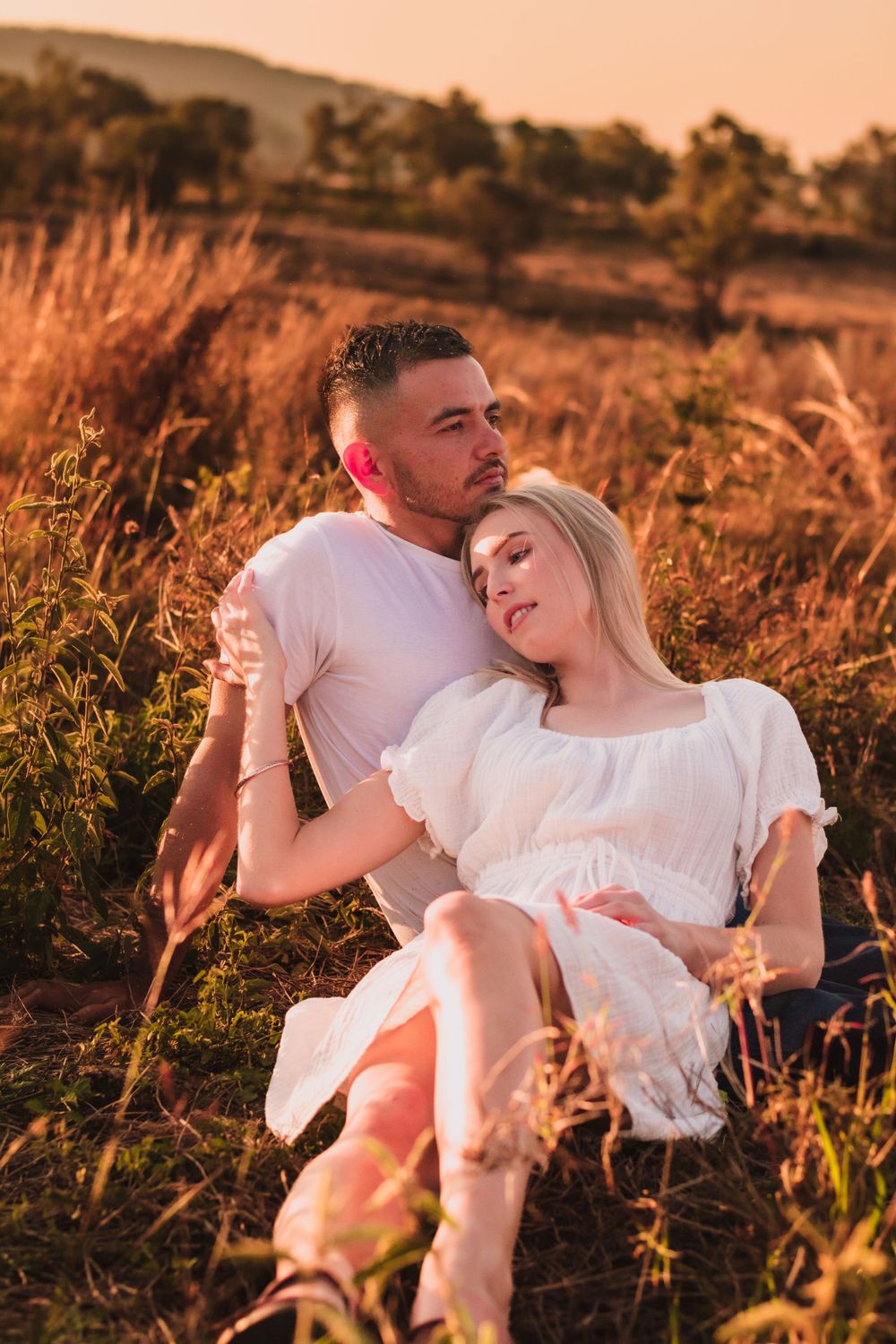 Couple relaxes in tall grass during golden hour as sun sets over rolling hills.