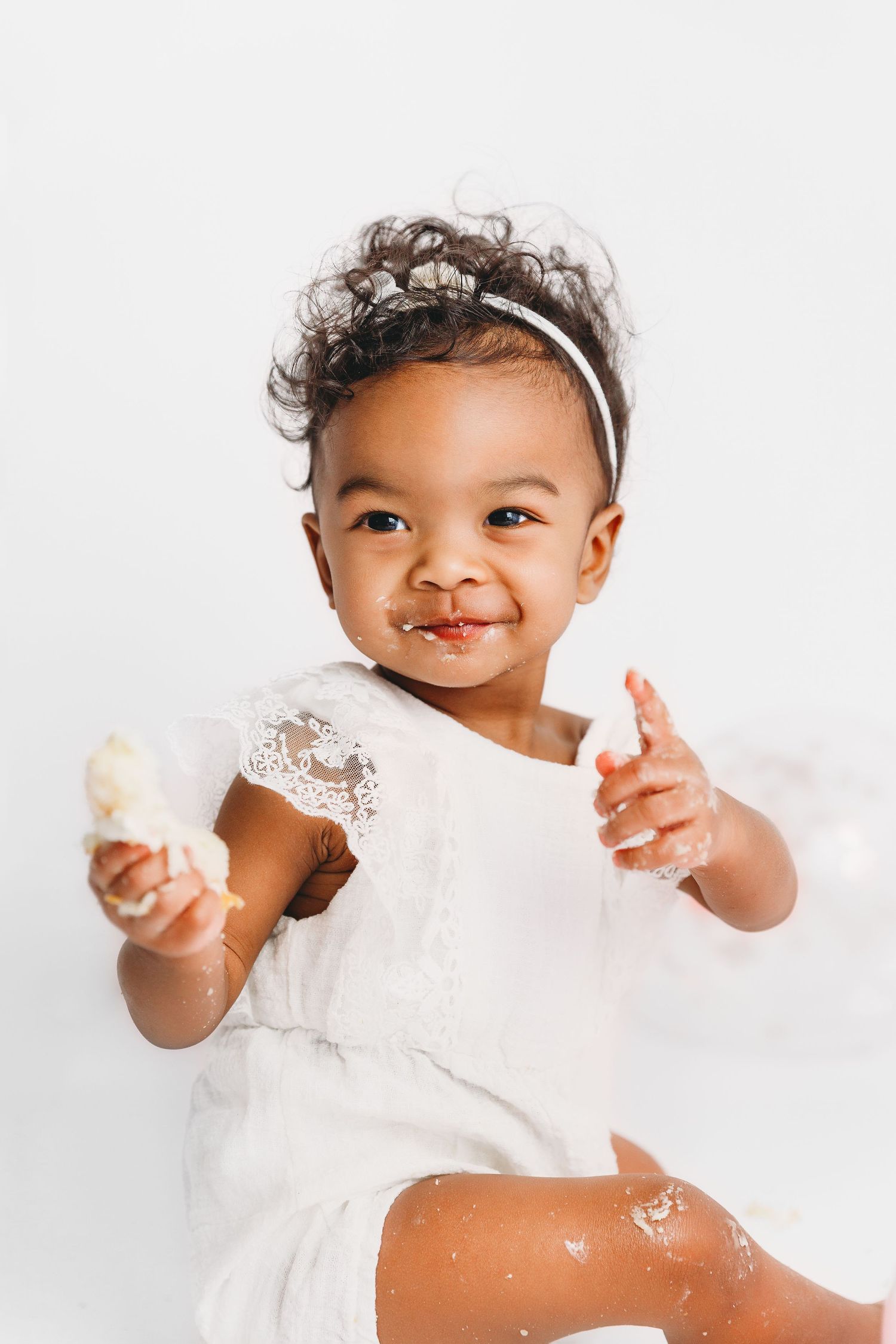 Baby with cake in hands and on face, smiling and pointing at the camera