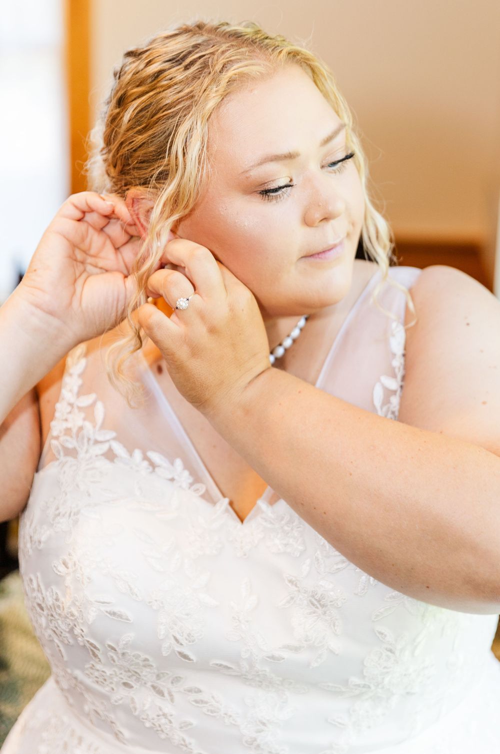 Bride in lace wedding dress fastens earrings while getting ready for her wedding ceremony.