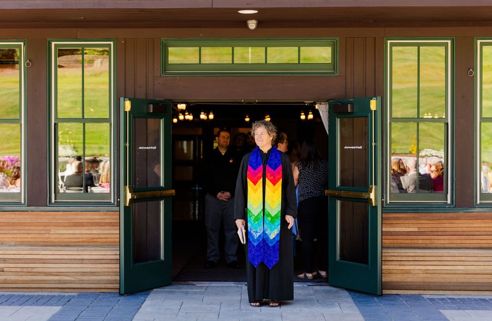 Officiant in rainbow stole stands in doorway of rustic venue during wedding ceremony.