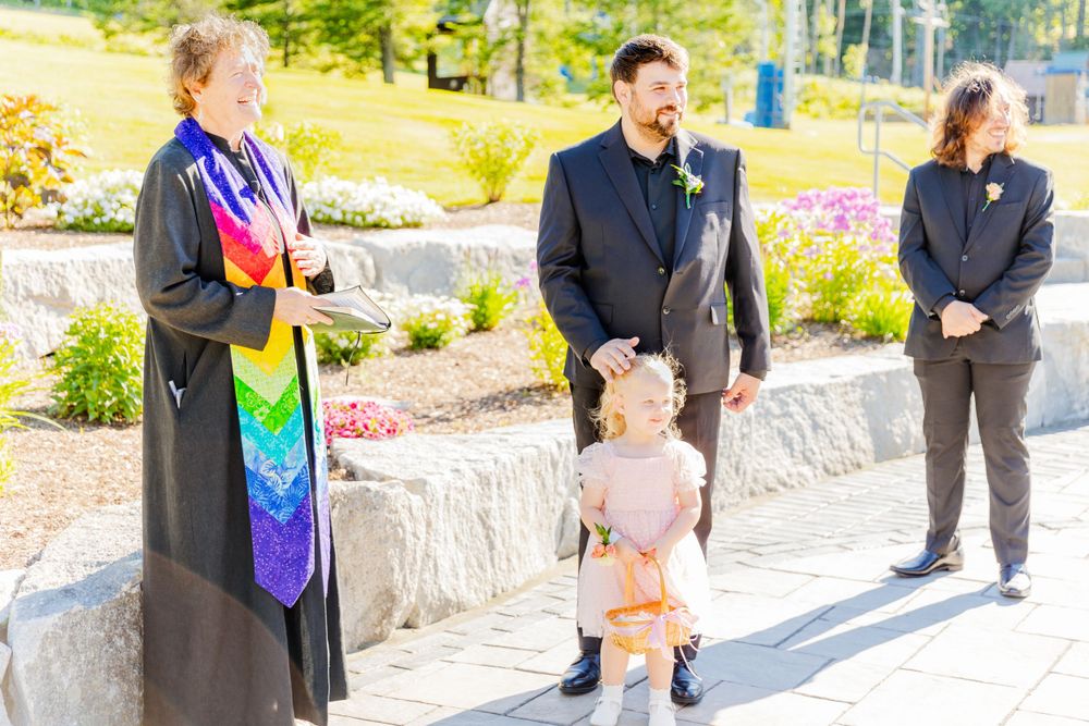 Outdoor wedding ceremony with officiant in rainbow stole and attendees on stone patio.
