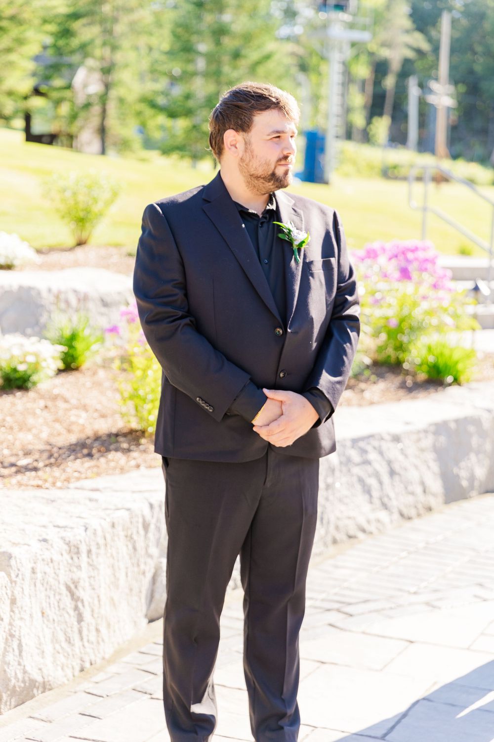 Groom in black suit stands on stone patio during outdoor wedding ceremony.