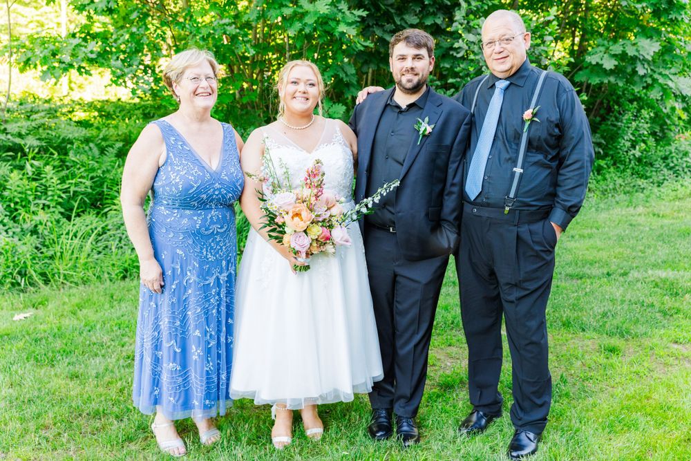 A wedding party poses outdoors in formal attire with the bride holding a colorful bouquet against lush greenery.