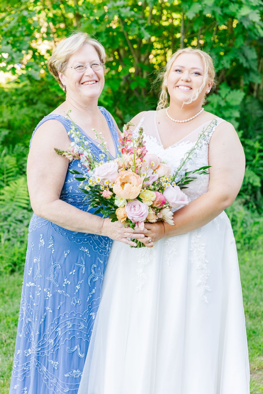 Two attendees in blue and white dresses share a moment with a pastel floral bouquet outdoors.