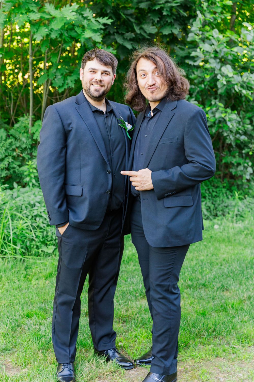 Two attendees in dark suits pose together on grass with green foliage in background.