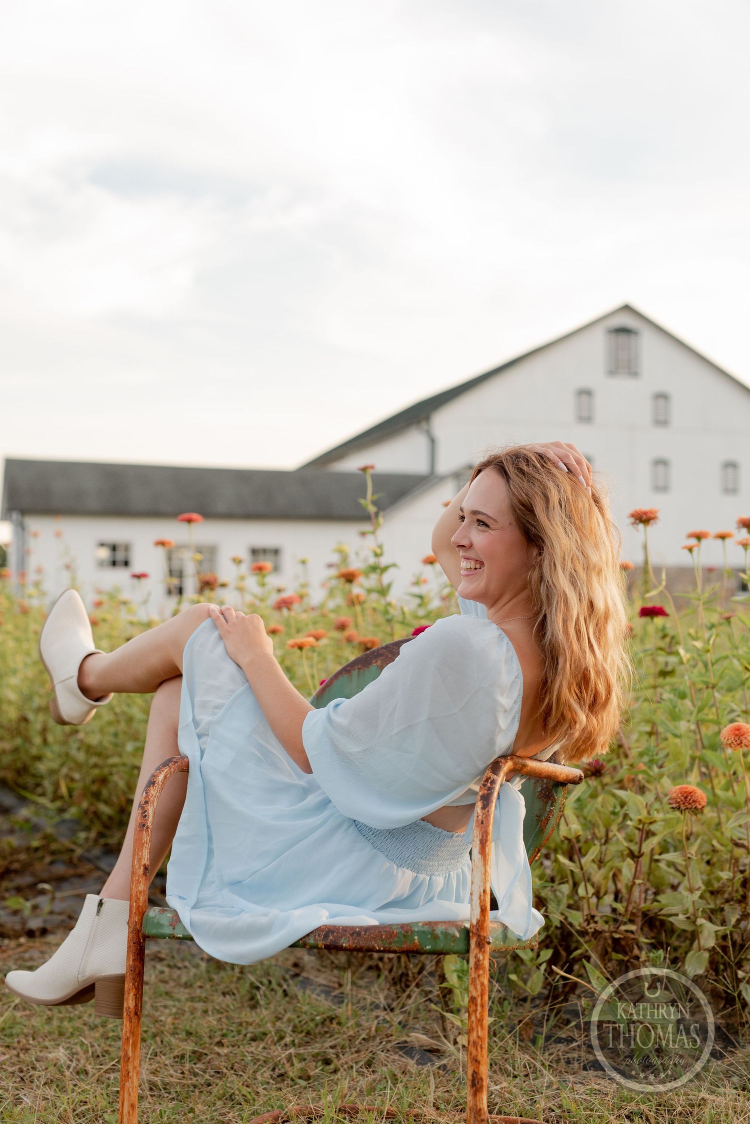 A playful pose in a light blue dress while sitting on a wooden chair among garden flowers.