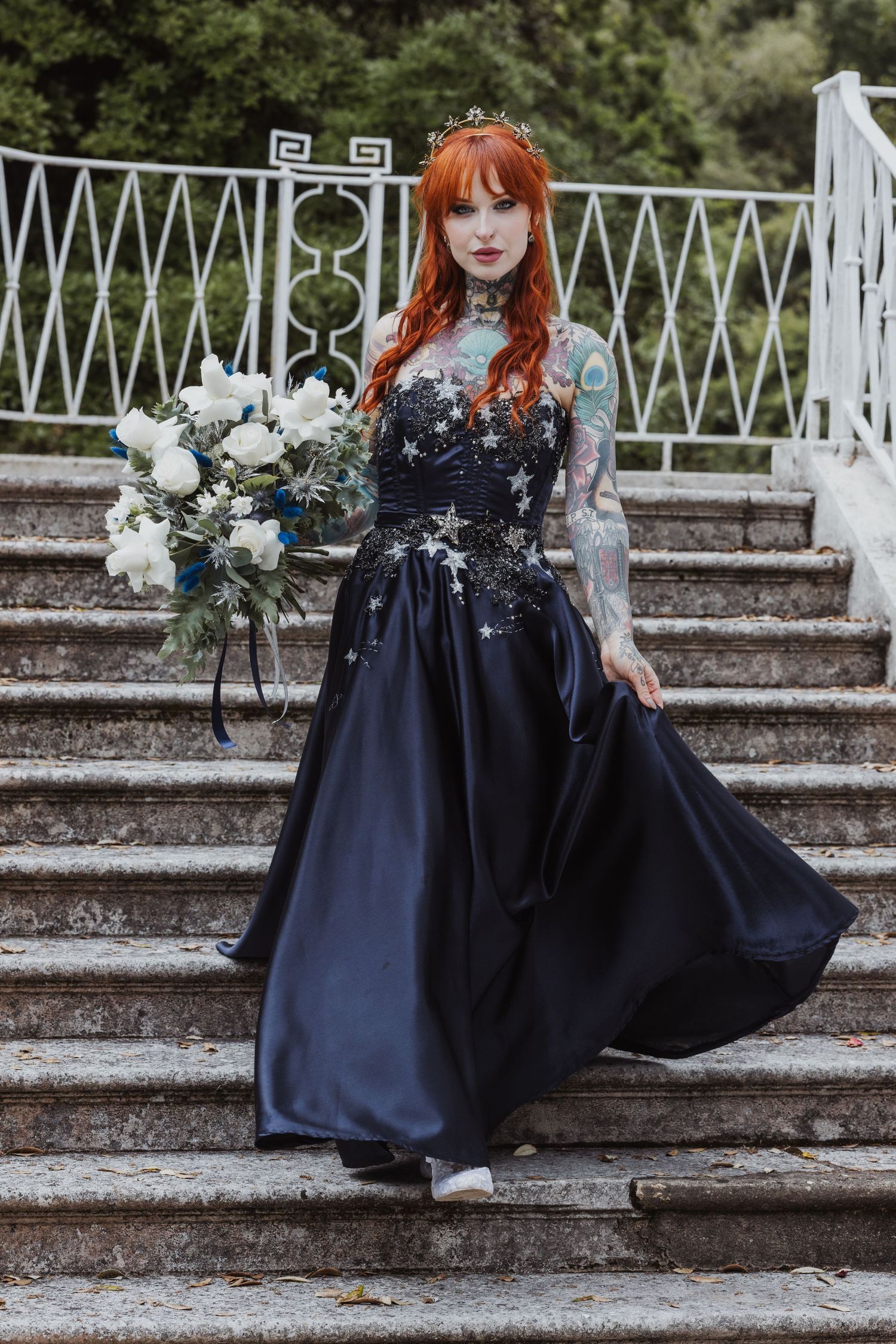 Bride in a navy gown with star embellishments, holding a white and blue bouquet, standing on stairs.