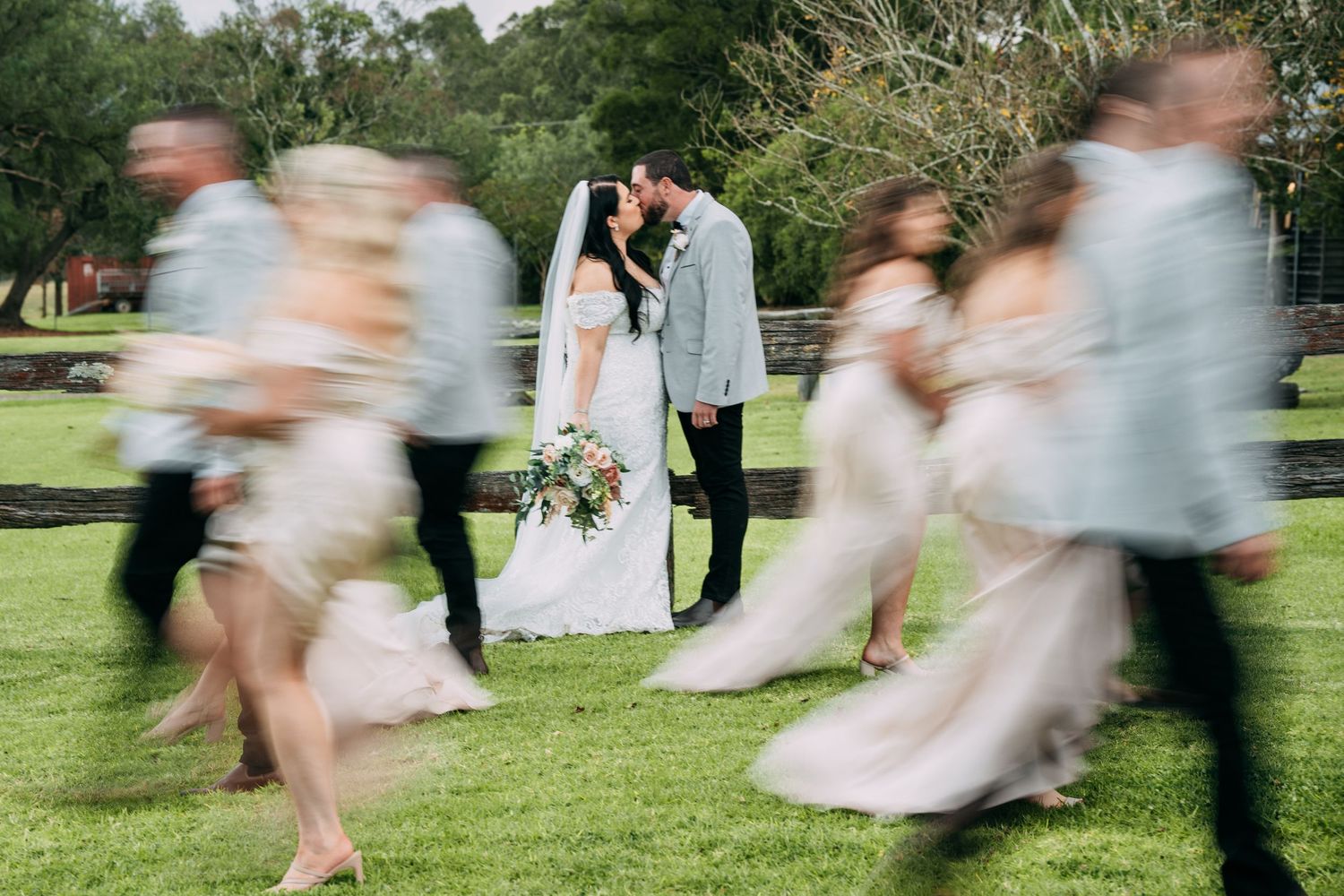 Motion-blurred wedding party dances around a couple on a green lawn.