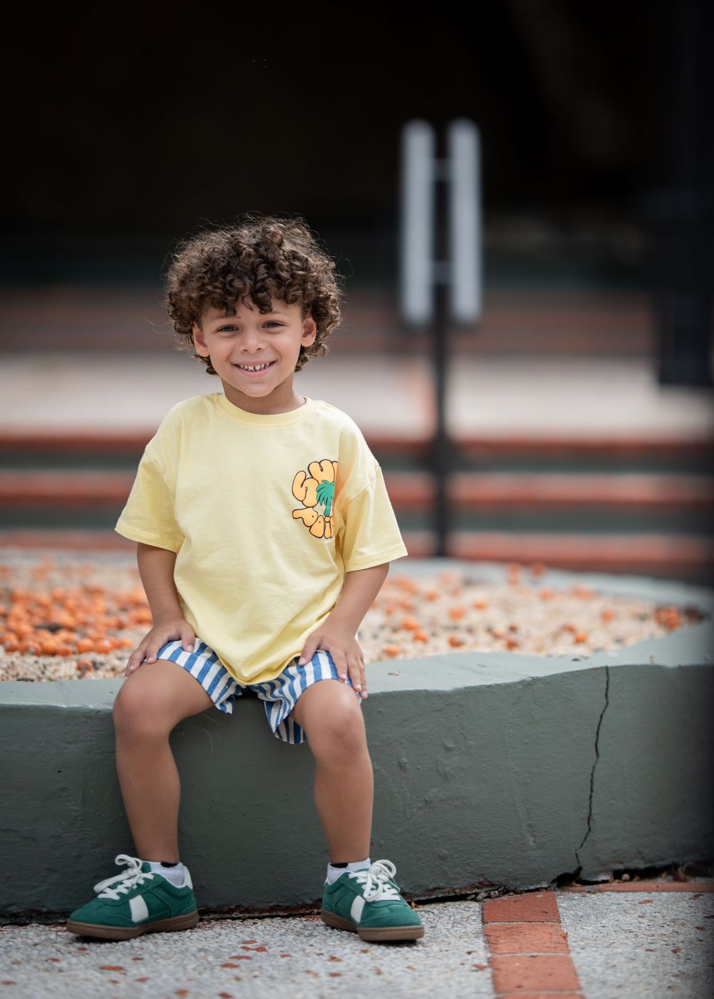 A child in yellow t-shirt and striped shorts sits on concrete steps outdoors.