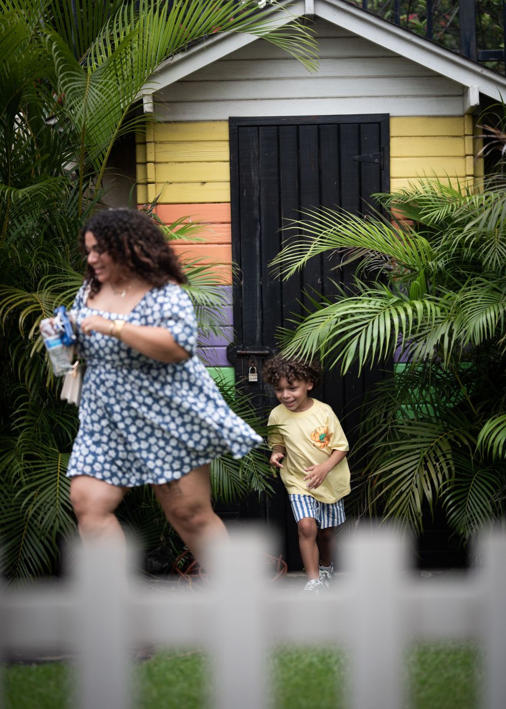 Two people walking past a yellow house with palm trees and black shutters.
