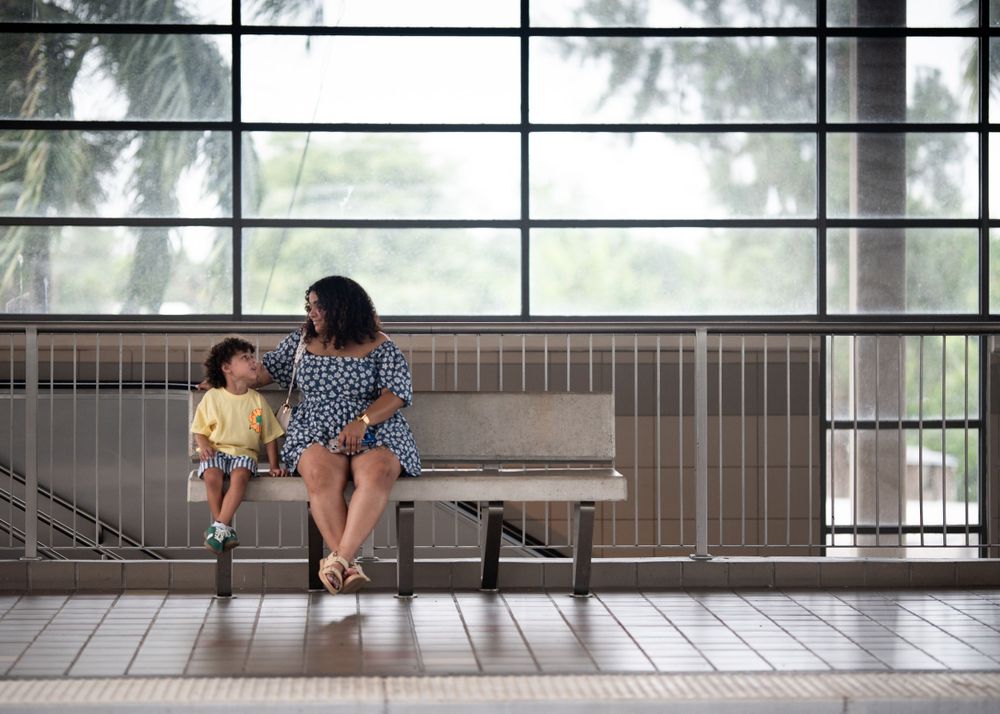 Two people sitting on a bench by large windows on a rainy day.