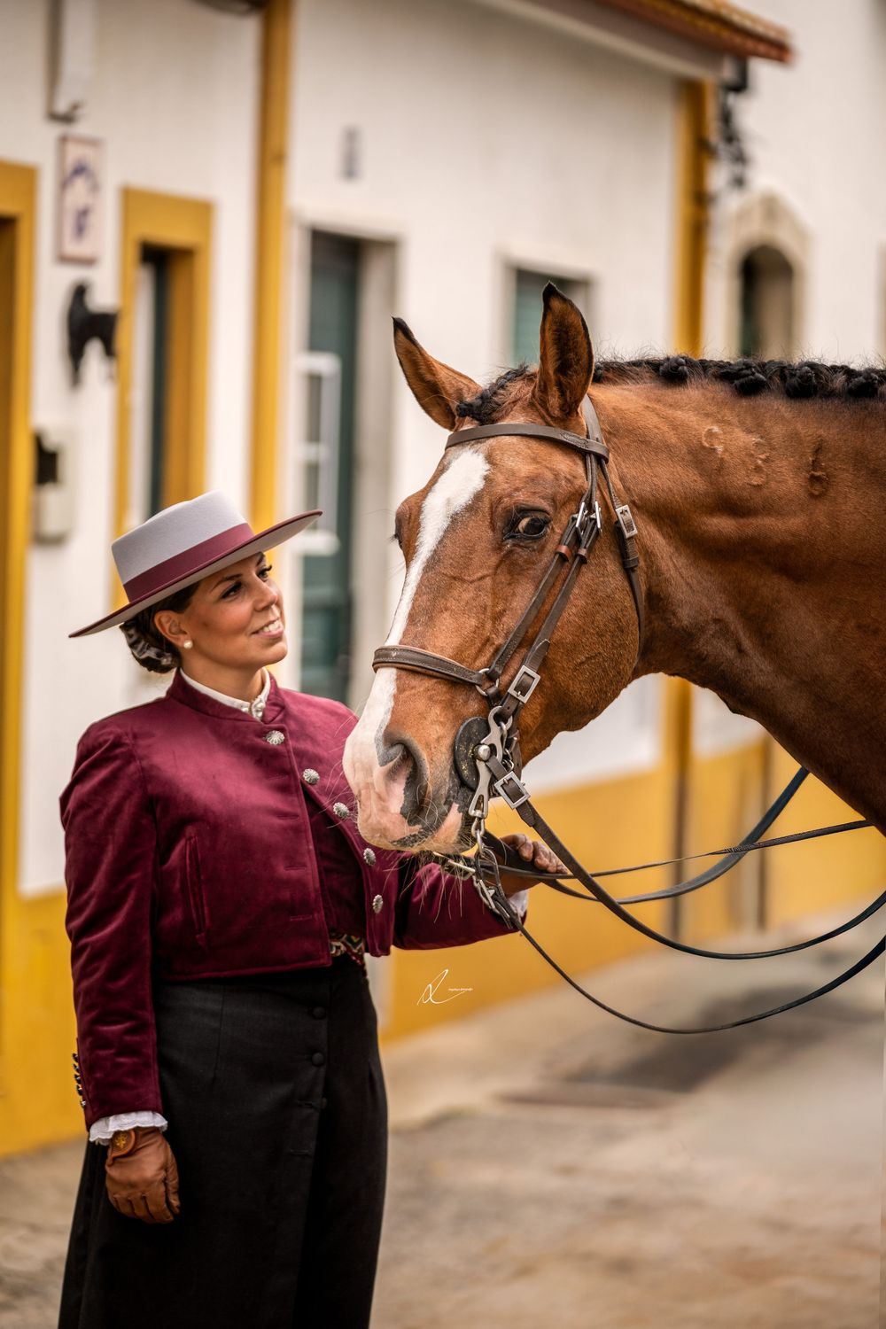 A rider in burgundy shirt and black pants stands with a brown horse against a white and yellow building exterior.