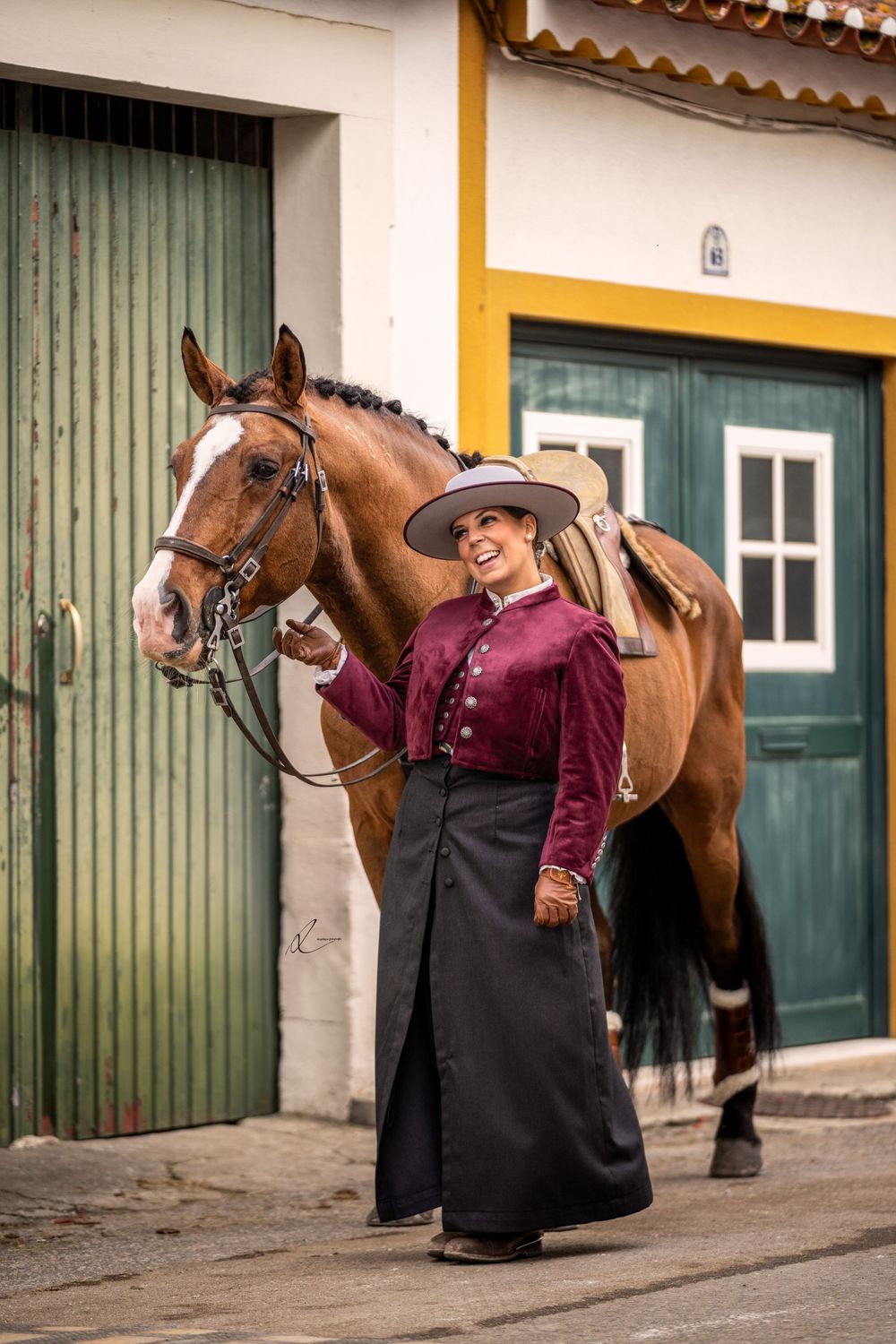 Traditional equestrian rider in burgundy velvet and black skirt stands with brown saddled horse by rustic stable doors.