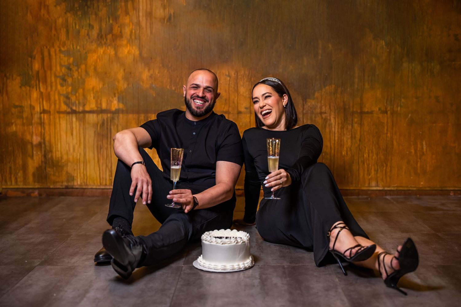 A couple celebrates with champagne and cake against an industrial rustic backdrop.
