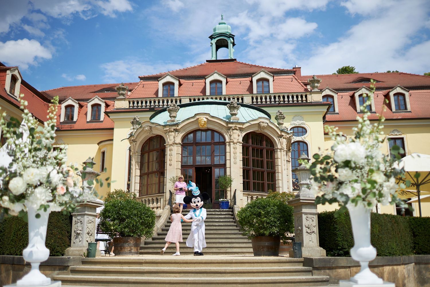 Beautiful wedding ceremony at a grand estate with white floral arrangements and architectural features set against blue skies.