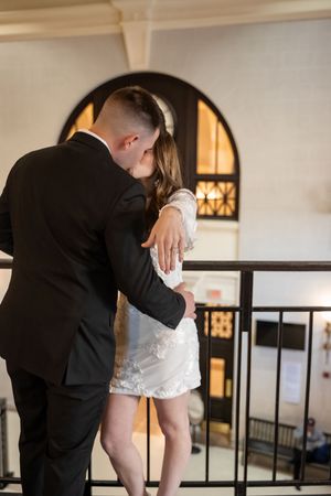 A couple in formal attire embraces intimately near an arched window during an indoor photoshoot.