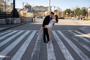 A couple shares a romantic dip kiss while standing on white crosswalk stripes in an urban setting during daytime.