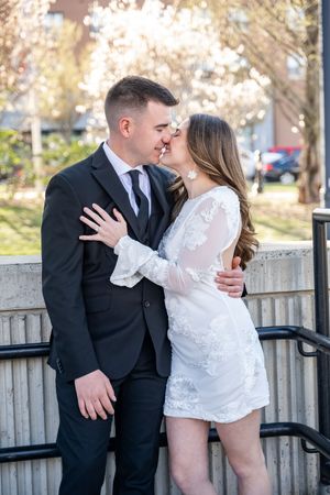 A romantic couple in formal attire embrace on concrete steps near flowering trees during a special occasion photoshoot.
