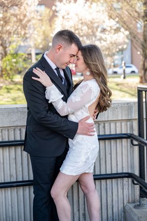 A romantic couple embraces outdoors in formal attire, with the bride wearing a short white lace dress and groom in black suit.