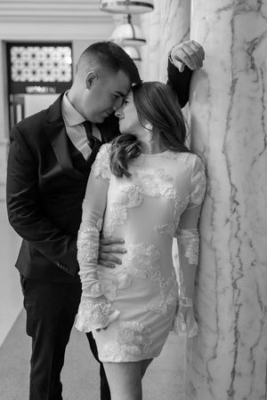 Romantic black and white portrait of a couple in formal attire embracing against a marble pillar.