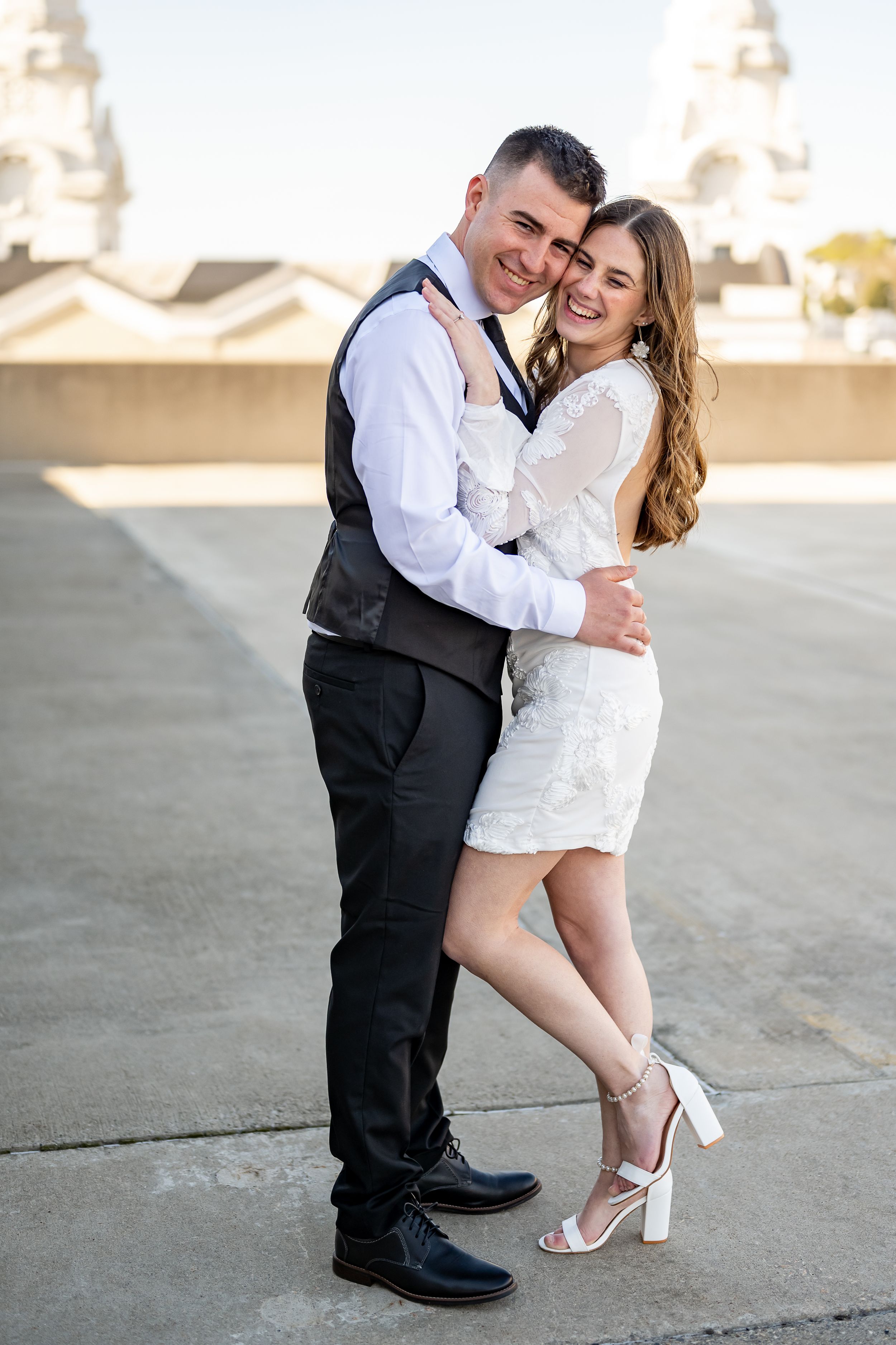A couple embraces and laughs together on a rooftop with architecture visible in the background.