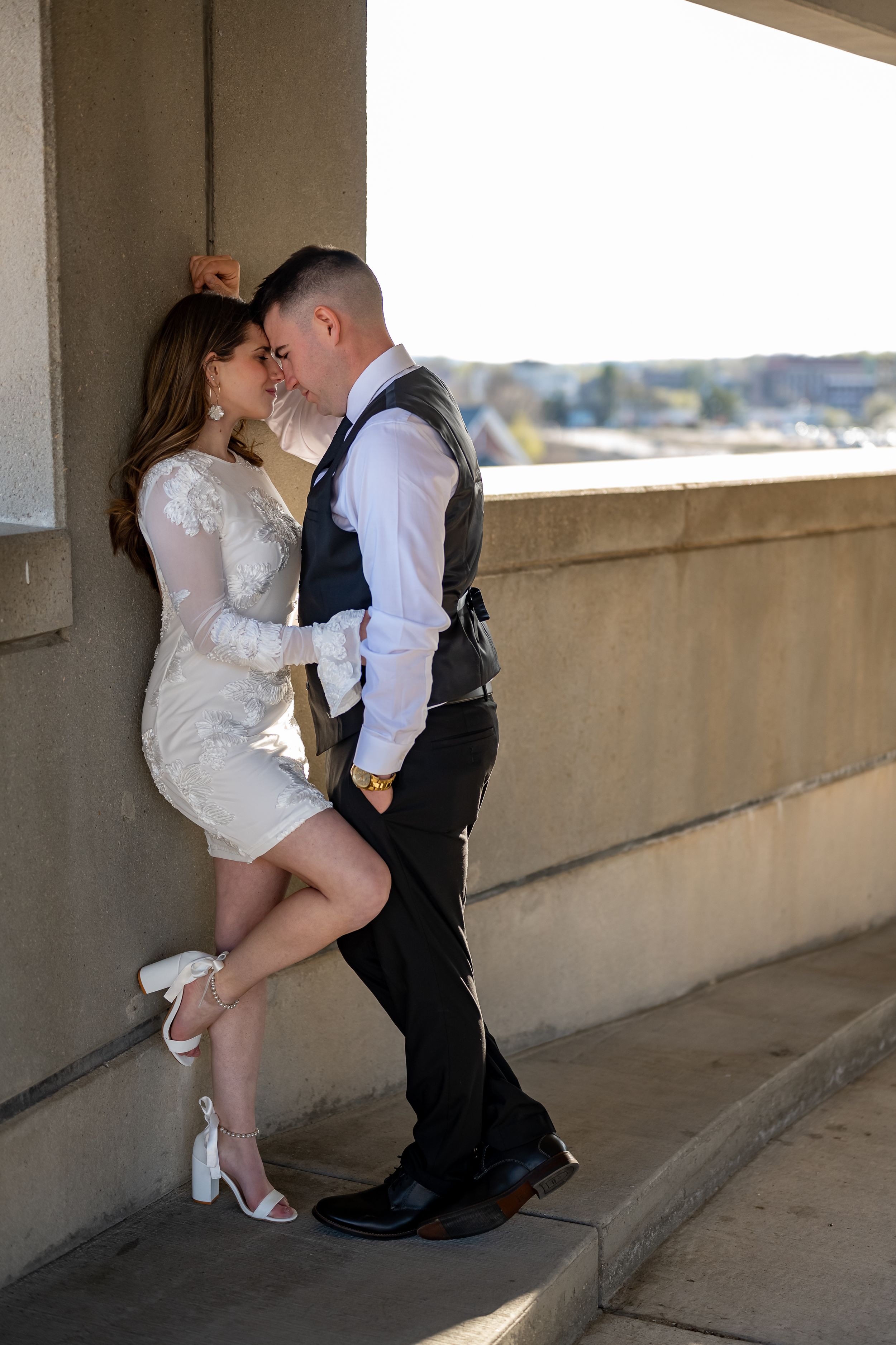 A romantic moment between a couple in wedding attire on a sunny outdoor balcony with white dress and formal vest.