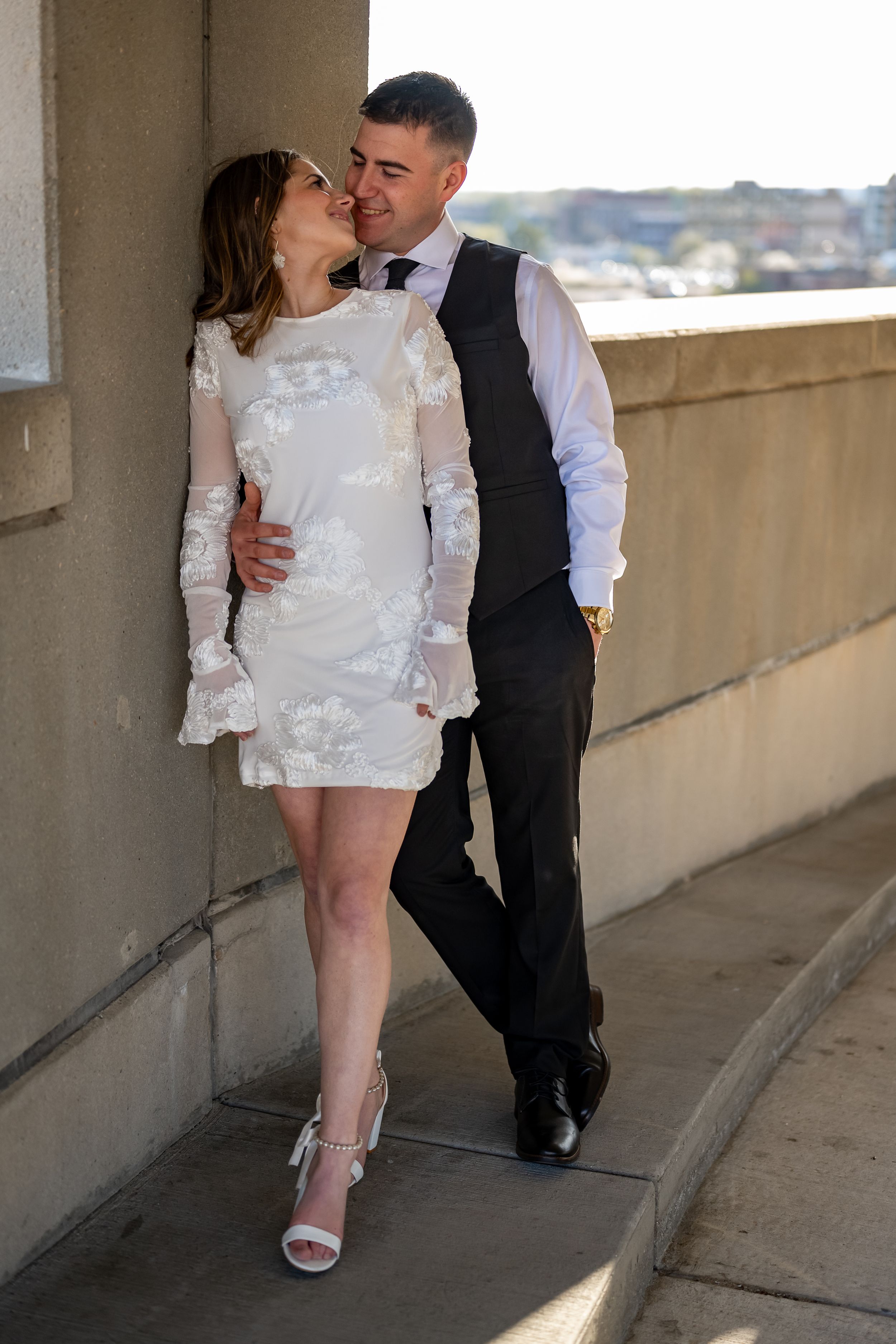 Couple in formal wear sharing an intimate moment on a balcony, with bride in short lace dress and groom in vest and tie.