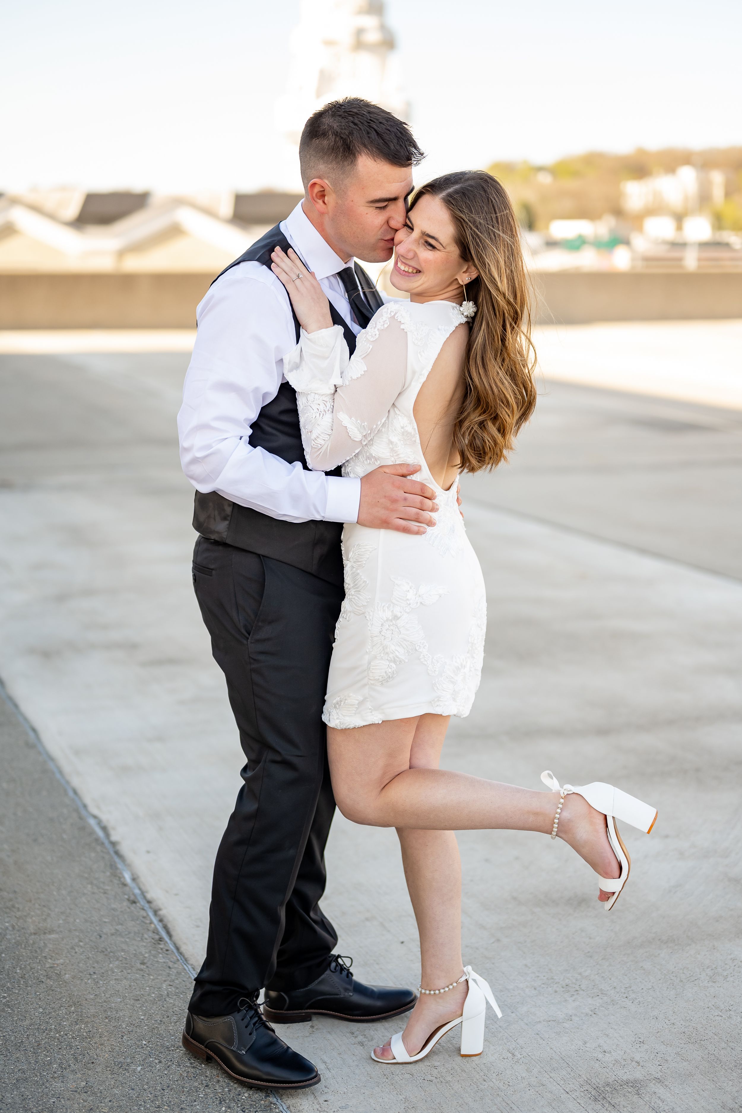 Romantic couple in formal attire embrace on rooftop at sunset, woman in white dress and man in dress shirt and vest.