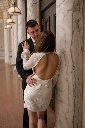 Intimate moment between couple in formal wear, with bride in backless lace mini dress against marble columns.