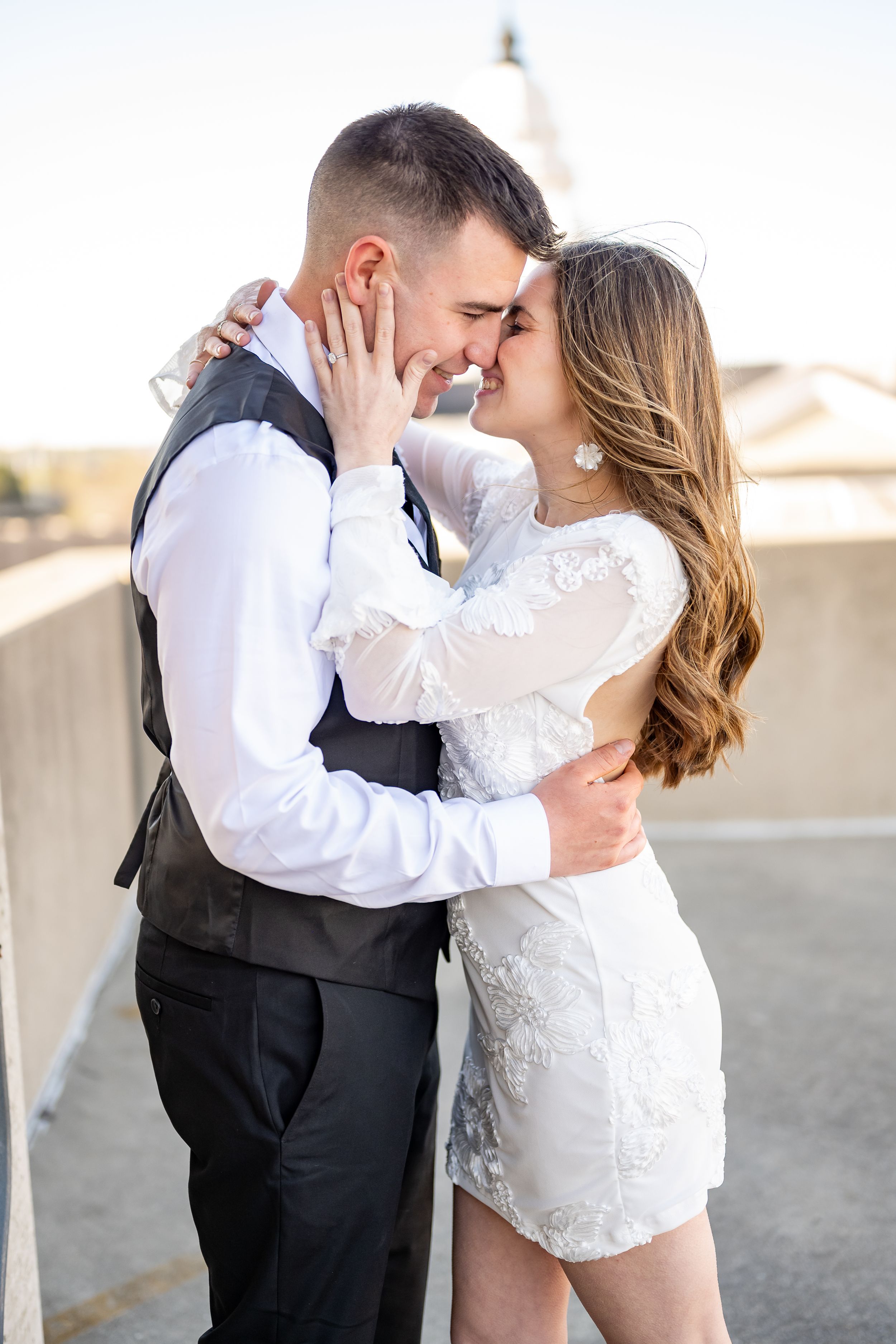 A romantic couple embraces and shares a kiss on a rooftop, with the bride wearing a short lace dress and groom in vest attire.
