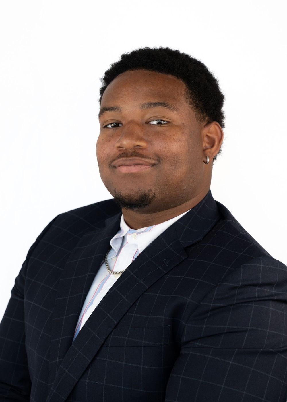 Professional headshot of a young man in a navy plaid blazer smiling confidently against white background.