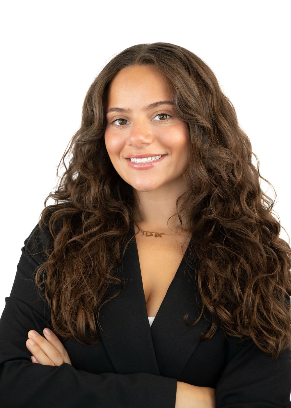 Professional headshot of a young woman with long curly brown hair in a black blazer, arms crossed.